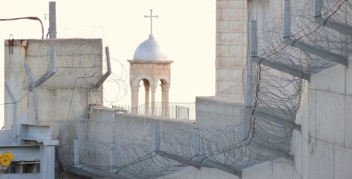 Jerusalem_Mount_of_Olives_Santa_Marta_Lazarus_church_above_the_wall A igreja de São Lázaro, no Monte das Oliveiras, construída no lado cisjordano, vê-se por trás do muro. Abed Salama vive do lado do muro em que todas as coisas se complicam, em que percorrer escassas centenas é um calvário moderno de ódio, racismo ou a fria burocracia. Foto © Utilisateur:Djampa - User:Djampa, CC BY-SA 4.0 , via Wikimedia Commons.