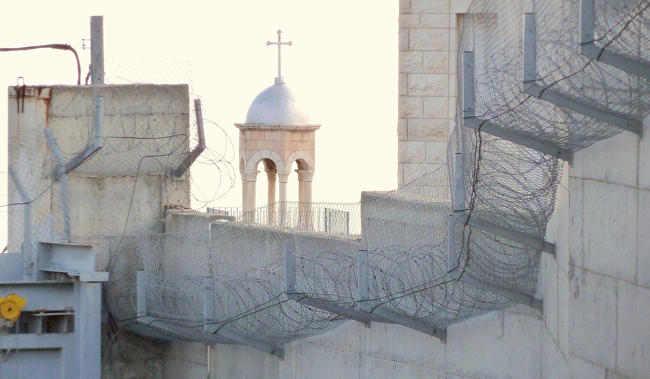 A igreja de São Lázaro, no Monte das Oliveiras, construída no lado cisjordano, vê-se por trás do muro. Abed Salama vive do lado do muro em que todas as coisas se complicam, em que percorrer escassas centenas é um calvário moderno de ódio, racismo ou a fria burocracia. Foto © Utilisateur:Djampa - User:Djampa, CC BY-SA 4.0 <https://creativecommons.org/licenses/by-sa/4.0>, via Wikimedia Commons.