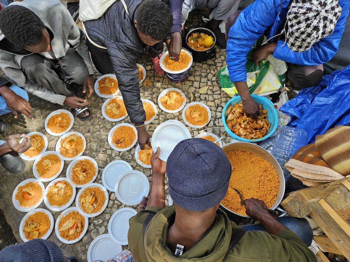 Pelas duas da tarde, o almoço está pronto. Num ápice, carregada por dois jovens, a pesada panela sobe os degraus do centro social para o clarão e o calor da rua. Foto © Cozinha Migrante dos Anjos.